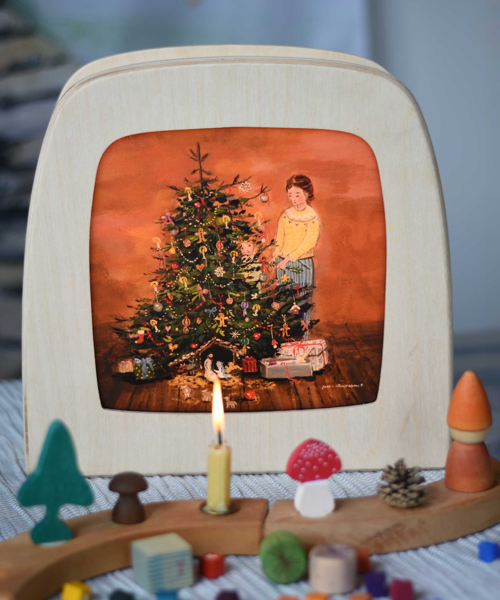 A Toverlux Christmas Tree Traditions Magic Lamp silhouette by artist Gwen van Knippenberg placed in a magic lamp on a table with a decorated Grimm's celebration ring in front of it. A watercolour and pencil style illustration of a woman and child standing by a decorated Christmas tree with candles on.