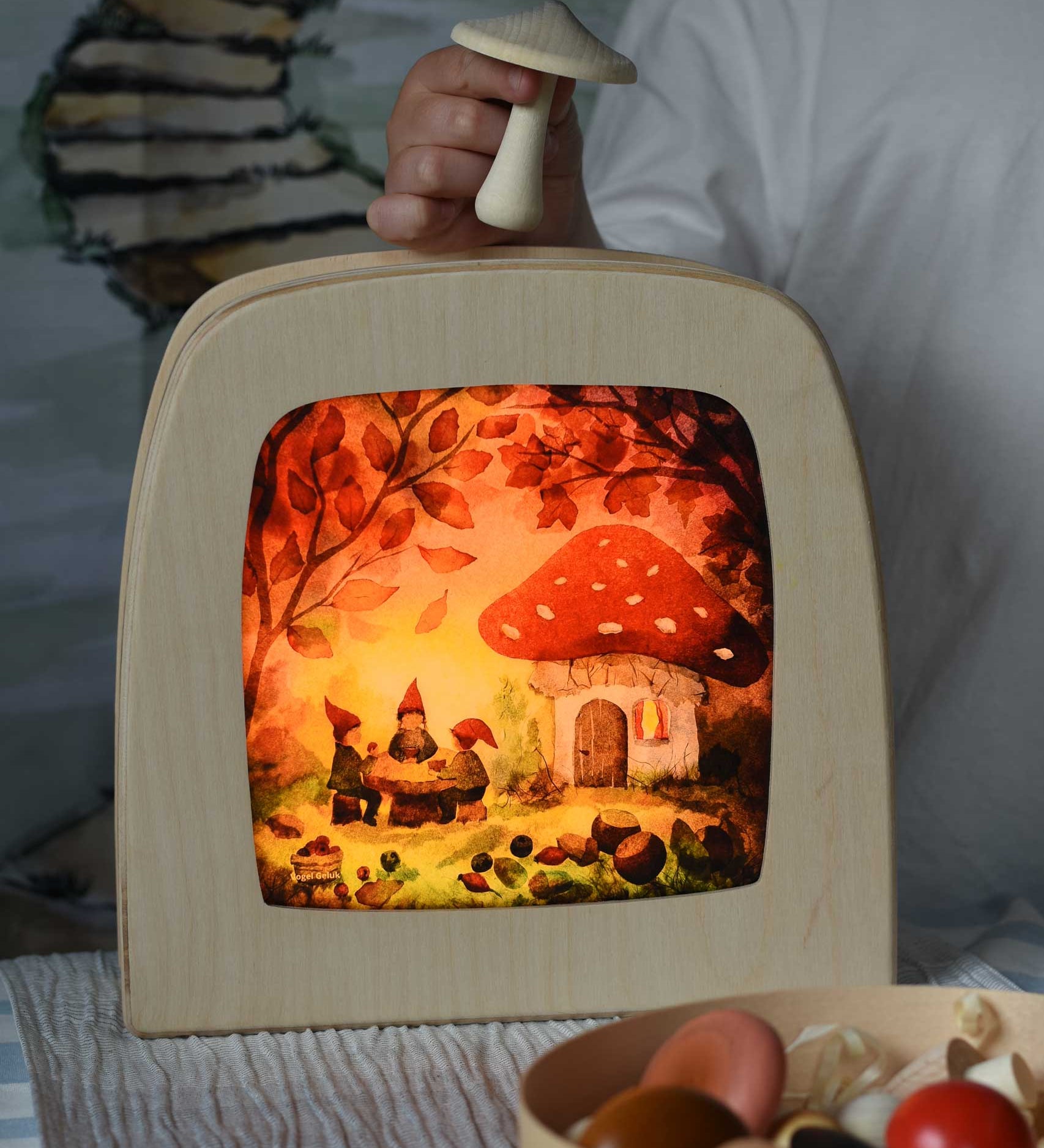 Toverlux magic lamp with a Autumn gnome toadstool picnic silhouette by artist Vogel Geluk. A watercolour style illustration of a toadstool house in a forest with a table with three gnomes sat around it. The silhouette has been placed in a magic lamp on a table. A child can be seen in the background placing Erzi mushrooms on top of the magic lamp.
 A range of Toverlux silhouettes, lamps and accessories are available at Babipur. 
