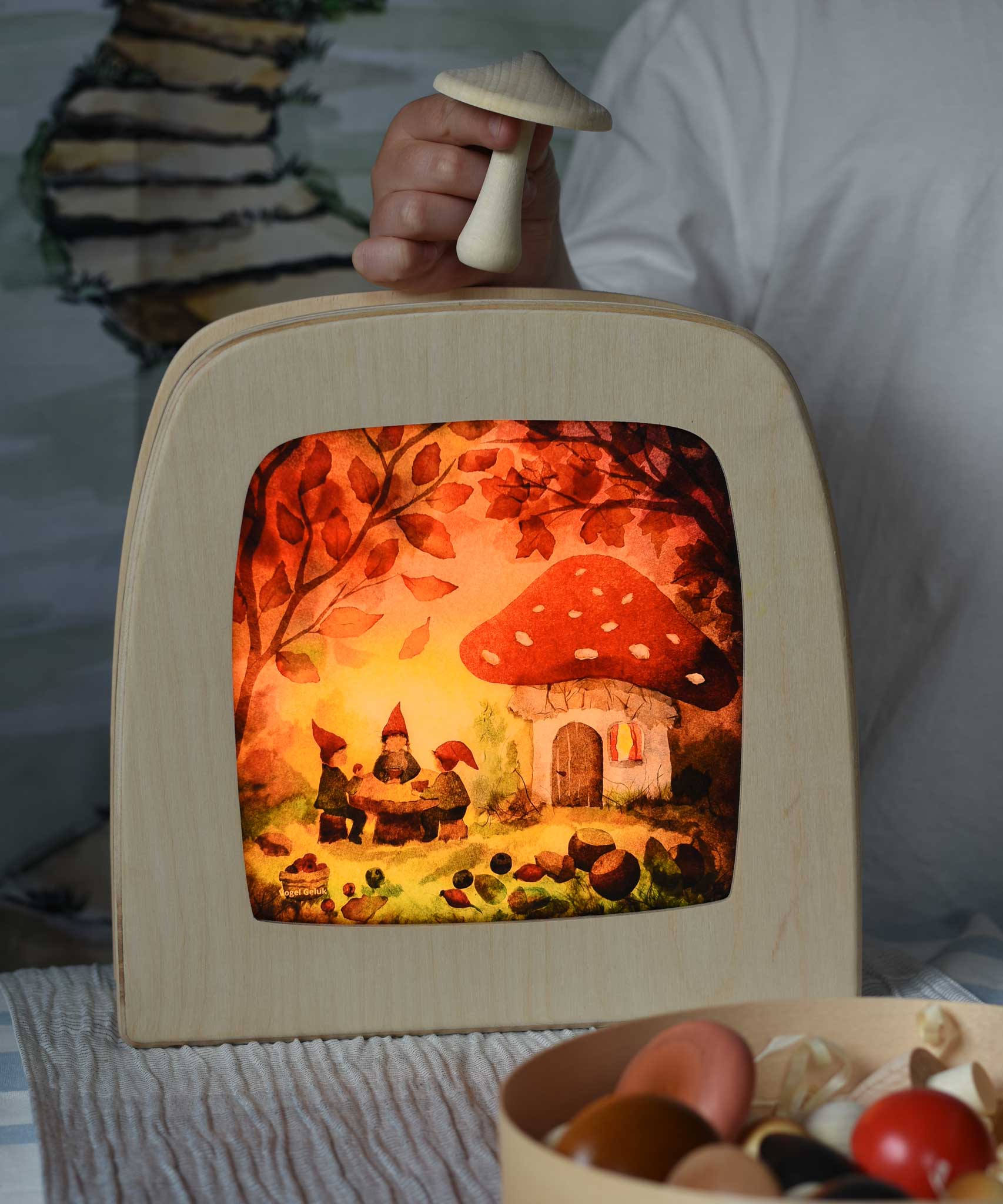 Toverlux magic lamp with a Autumn gnome toadstool picnic silhouette by artist Vogel Geluk. A watercolour style illustration of a toadstool house in a forest with a table with three gnomes sat around it. The silhouette has been placed in a magic lamp on a table. A child can be seen in the background placing Erzi mushrooms on top of the magic lamp.
 A range of Toverlux silhouettes, lamps and accessories are available at Babipur. 