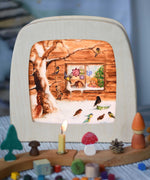 Toverlux Magic Lamp Silhouette - with Winter bird watching snow Artwork By Eentje Van Margo. A snow covered scene featuring birds feeding on a tree whilst small children look on from a window in the background. The silhouette has been placed in a magic lamp and placed on a table. There is a decorated Grimm's celebration ring in front of the lamp.
