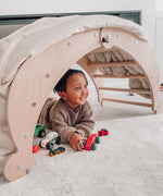 A close up of a Triclimb wooden Archi folding rocker climbing frame standing up. Showing a blanket on top acting as a den with a child playing underneath. 