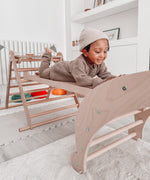 A Triclimb wooden Miri slide as a bridge in between a Archi folder rocker and Triclimb pikler climbing frame with a child lying on top of the slide in a playroom.