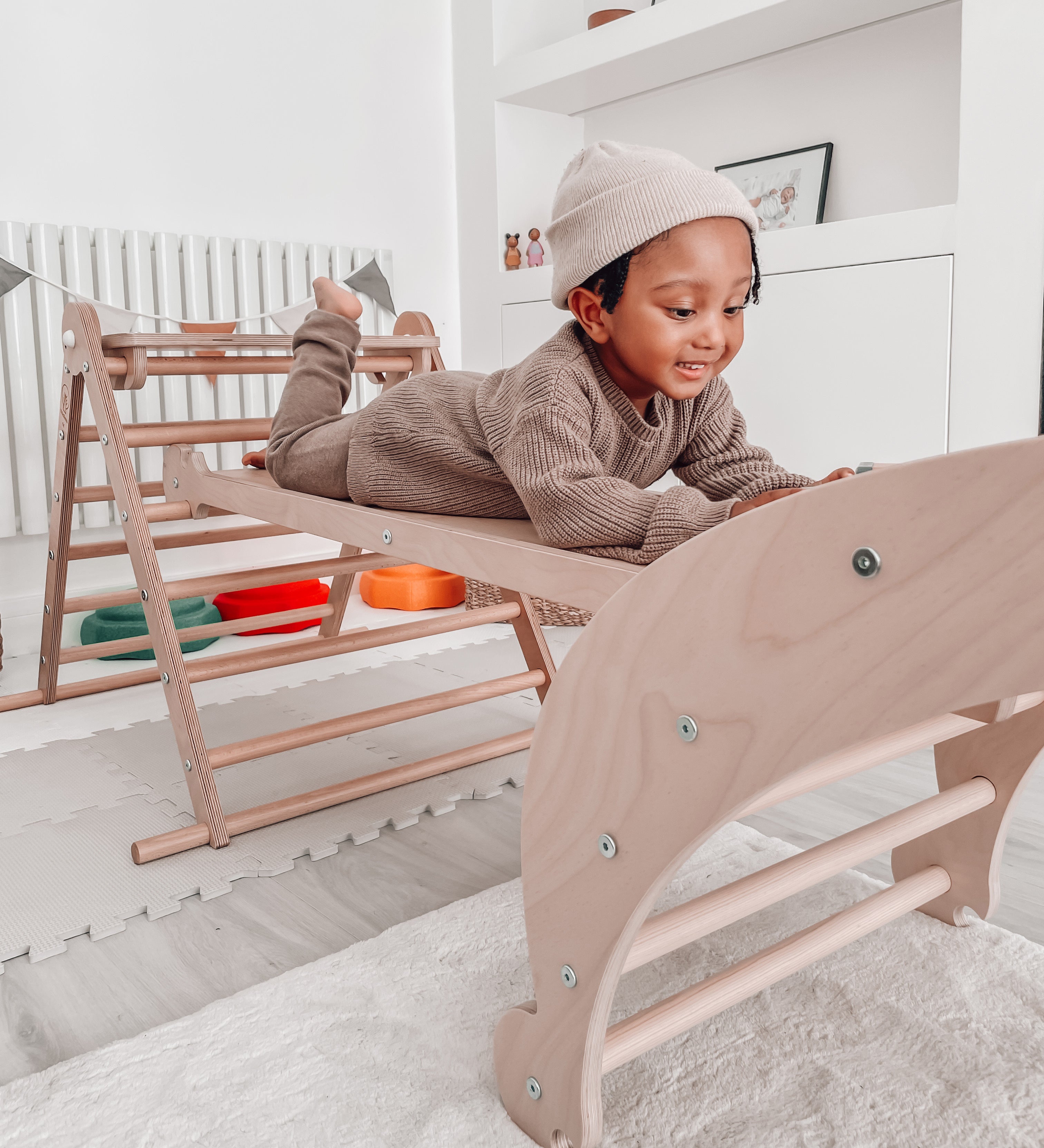 A Triclimb wooden Miri slide as a bridge in between a Archi folder rocker and Triclimb pikler climbing frame with a child lying on top of the slide in a playroom.