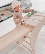 A child standing over the Triclimb wooden archi folding rocker climb frame and playing with various vehicles on the wooden slats with a Miri slide sitting on top of a rung acting as a bridge