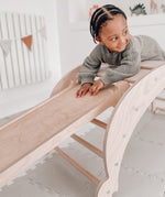 A child lying on top of the wooden slats on  the Triclimb wooden Archi folding rocker climbing frame with a Miri slide hooked on a rung 