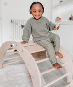 A child sitting on top of the slats of the Triclimb wooden Archi folding rocker climbing frame in a playroom