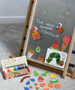 A Triclimb Biri placed on a rainbow Triclimb. A pack of Kitpas crayons have been placed on the floor in front of it. The board has been decorated with hand drawn food illustrations placed under Bajo gruffalo magnets and a caterpillar featured in the very hungry caterpillar book.  