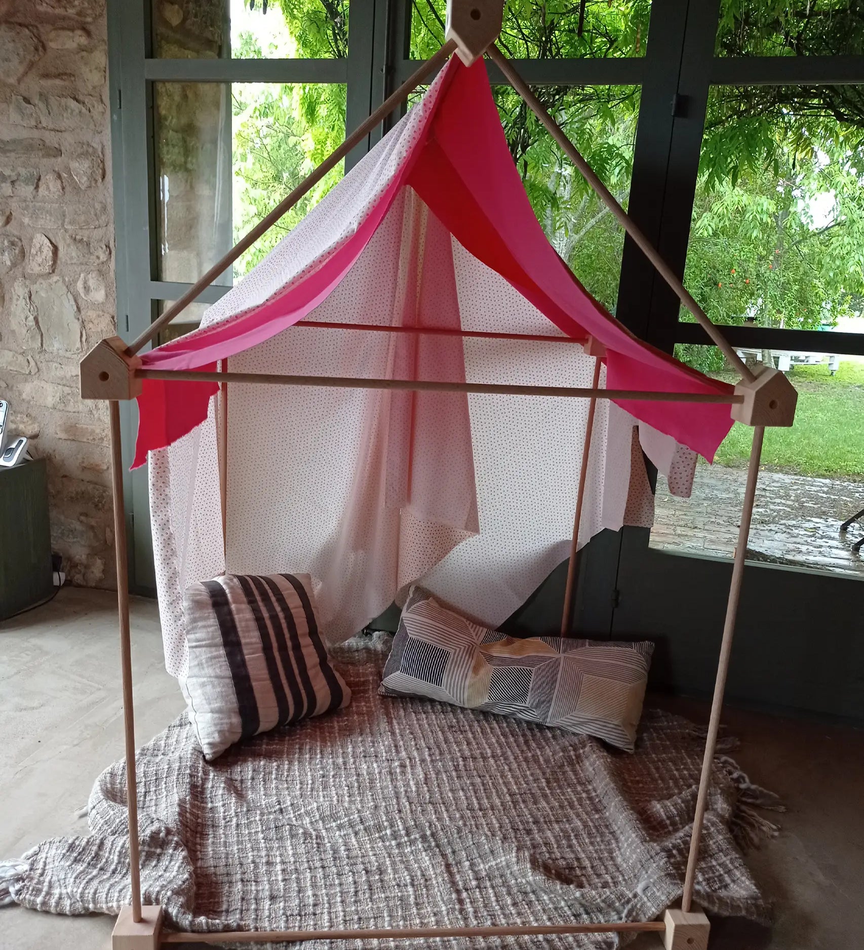 A cosy tent setup made from the Trigonos wooden big start set showing a pink cloth on top of the frame standing on a concrete surface