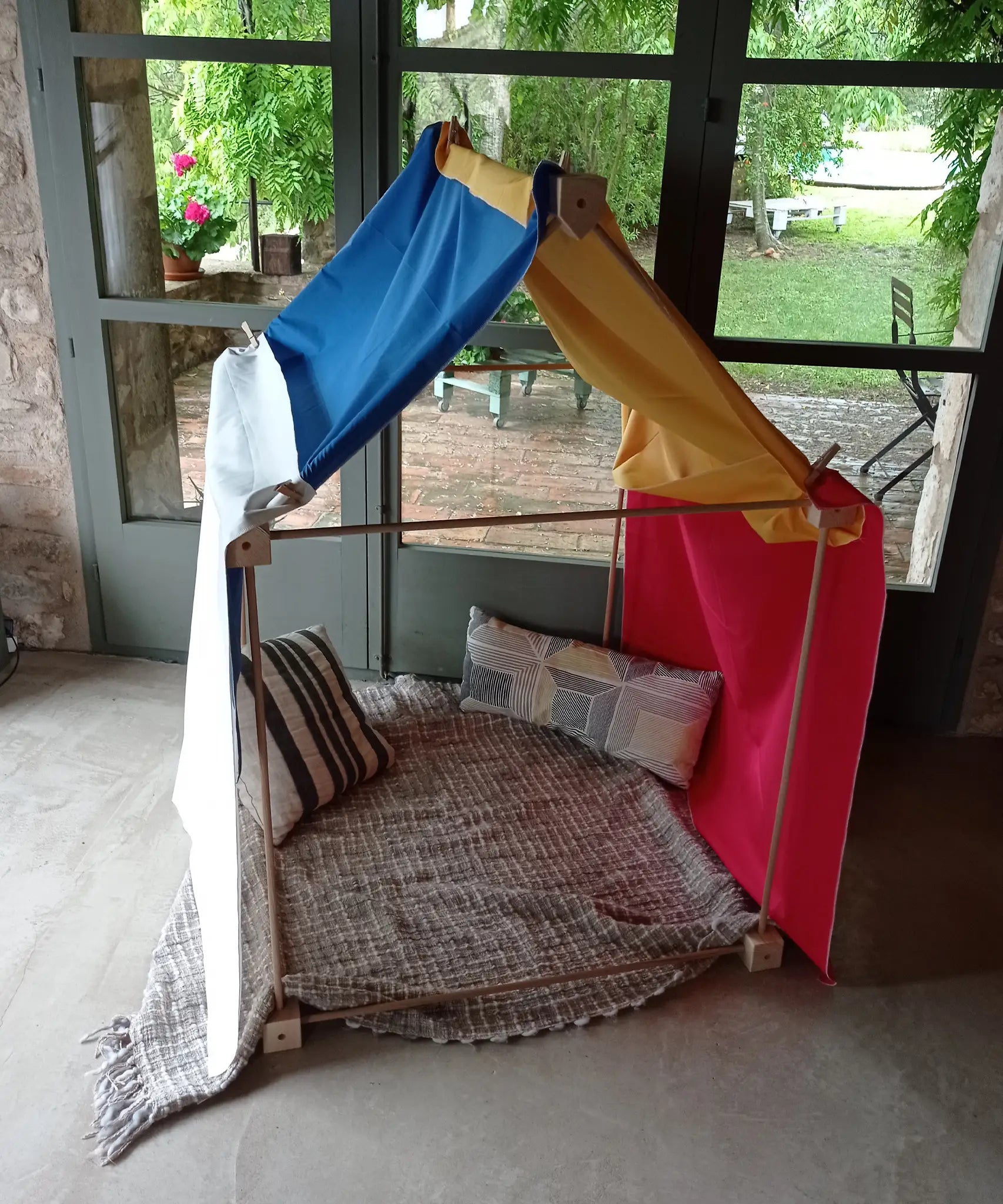 Tent made from the Trigonos wooden big start set showing coloured cloth on top of the frame with cushions and a blanket inside