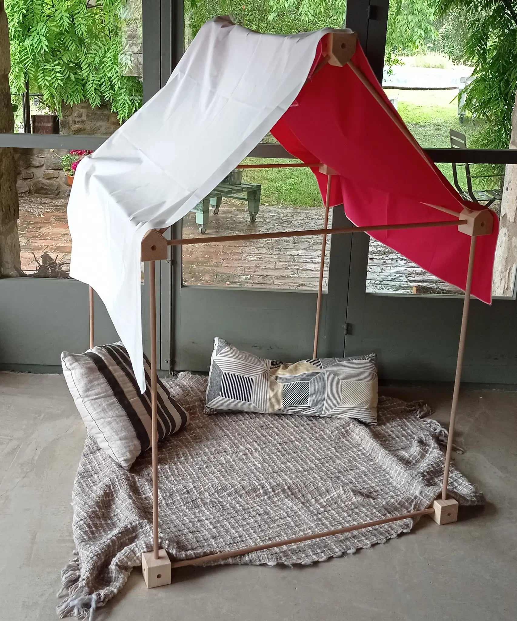 A tent made from the Trigonos wooden big start set showing a white cloth on top of the frame with cushions and a blanket inside