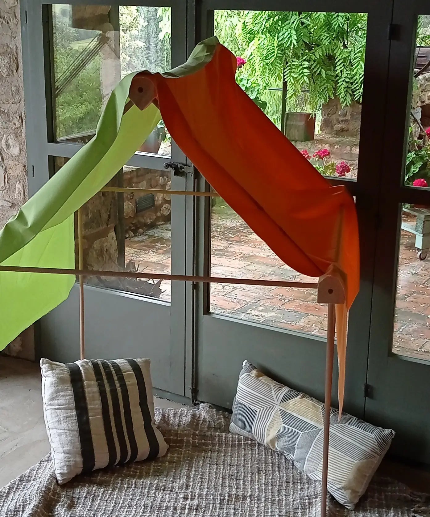 A tent setup made from the Trigonos wooden big start set showing a rainbow cloth on top of the frame and cushions inside