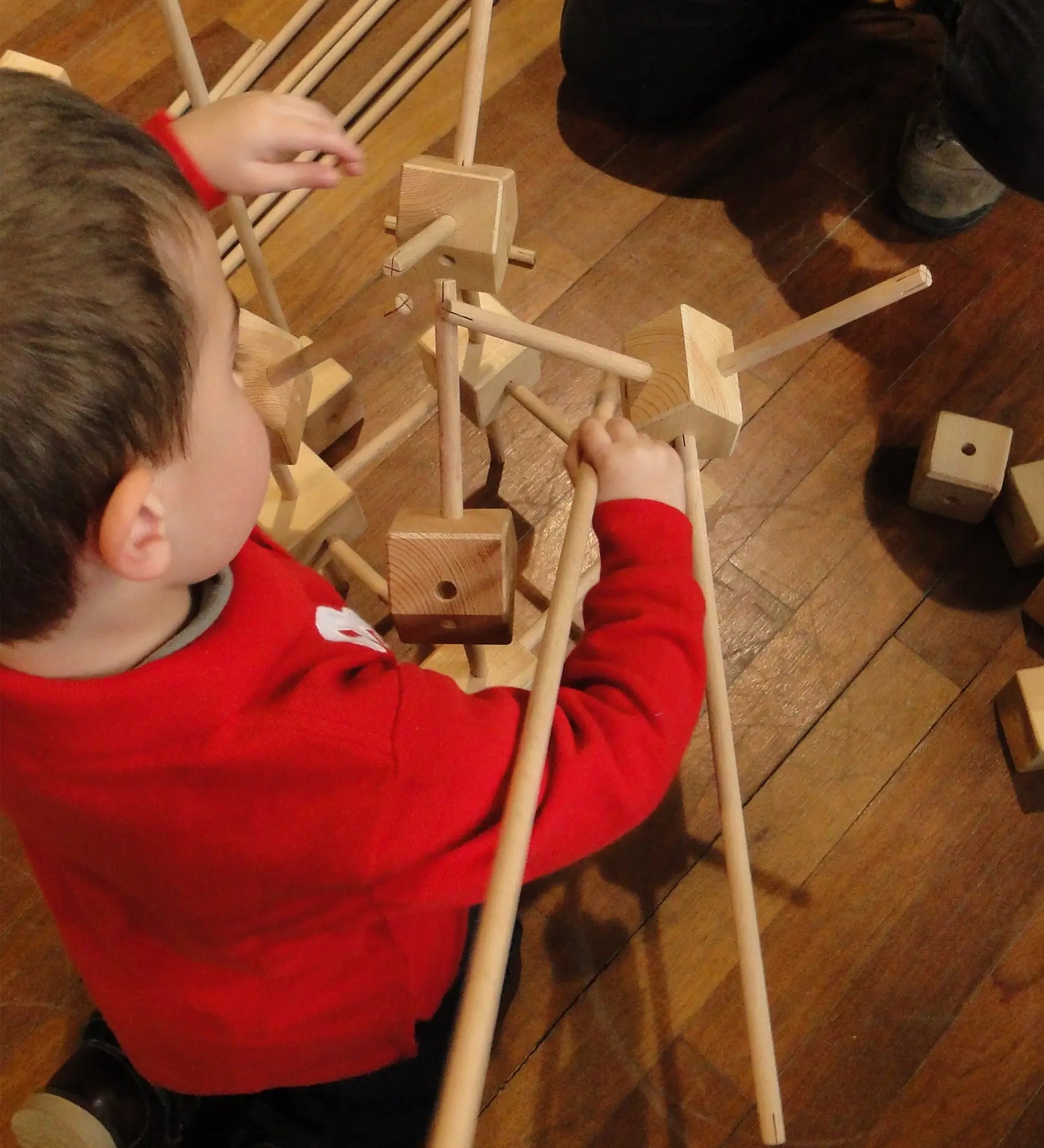 Child building with wooden poles and blocks from the Trigonos starter construction set on a wooden floor