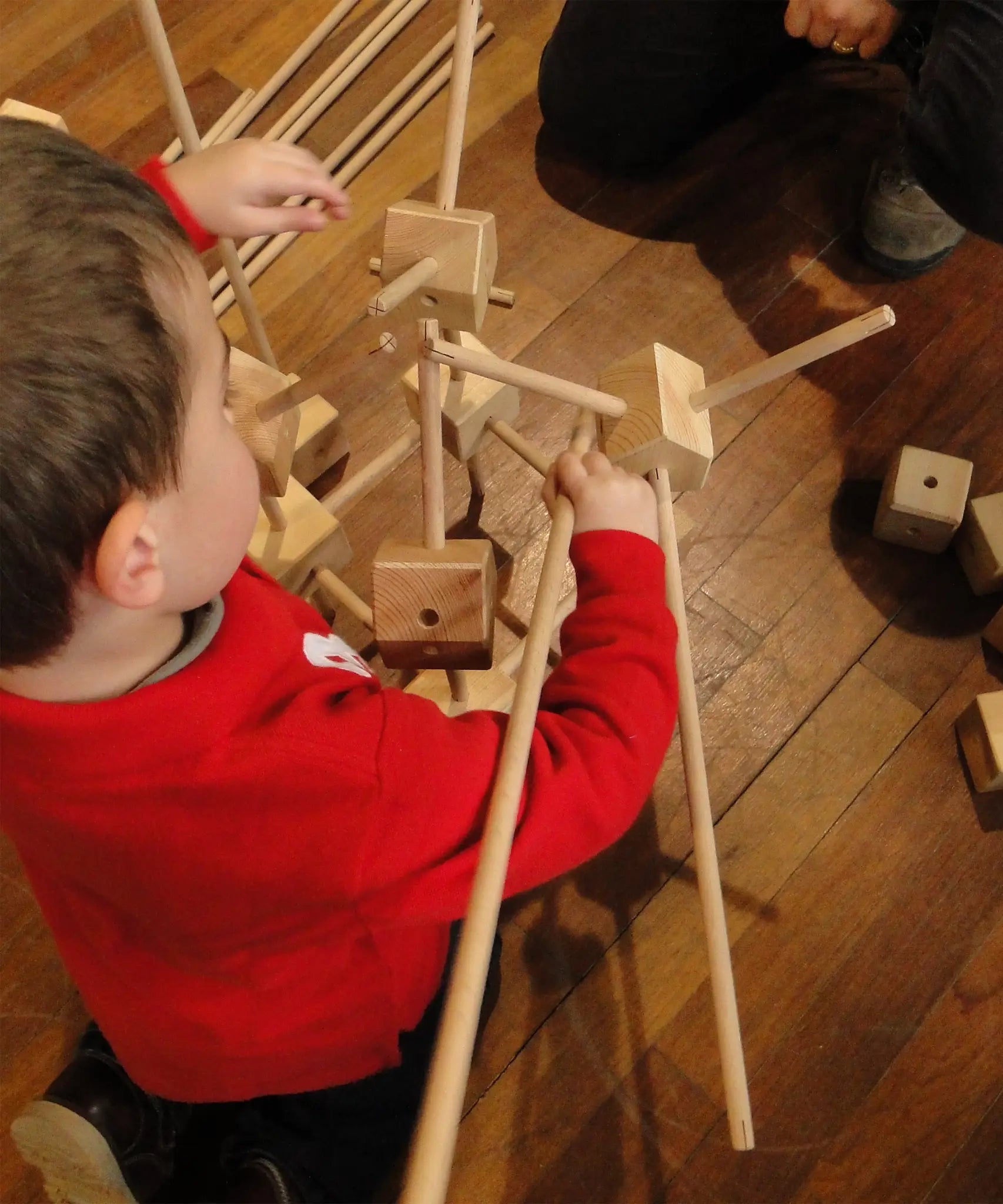 Child building with wooden poles and blocks from the Trigonos starter construction set on a wooden floor