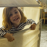 Child pulling a white cotton sheet down from the wooden frame made from the Trigonos kernel organic set