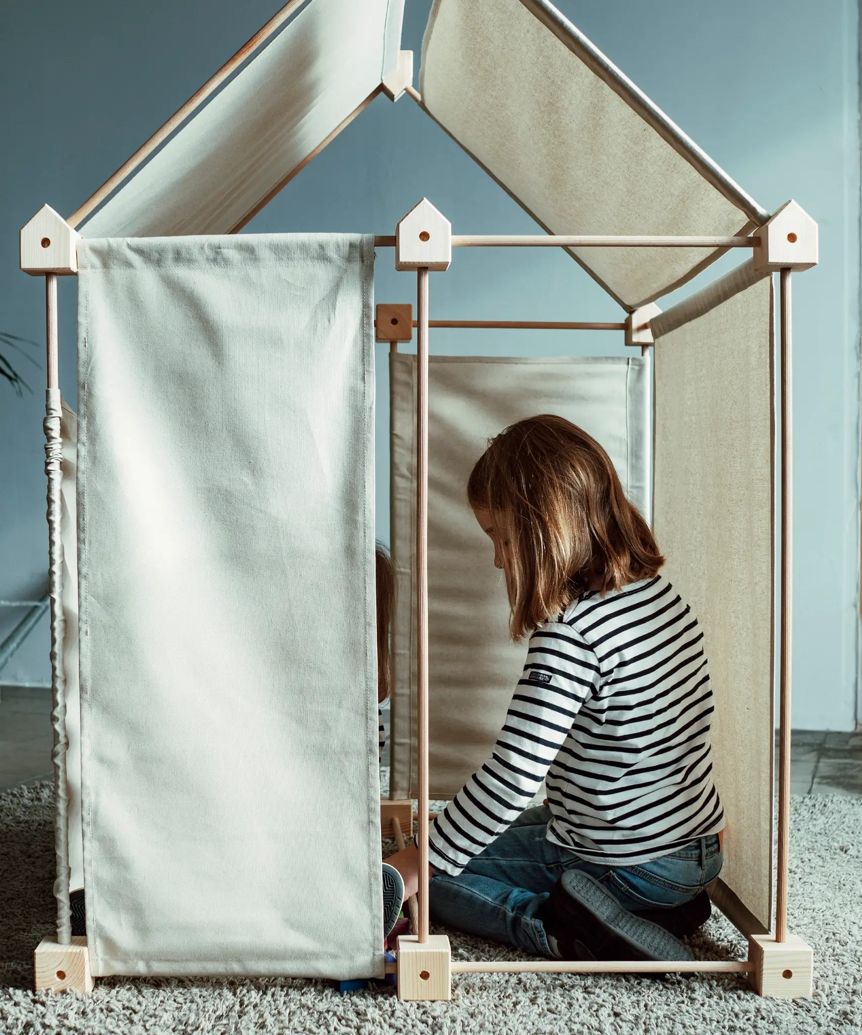 Child sitting inside a tent structure made from the Trigonos wooden kernel organic construction set with white cotton sheets on the roof and sides