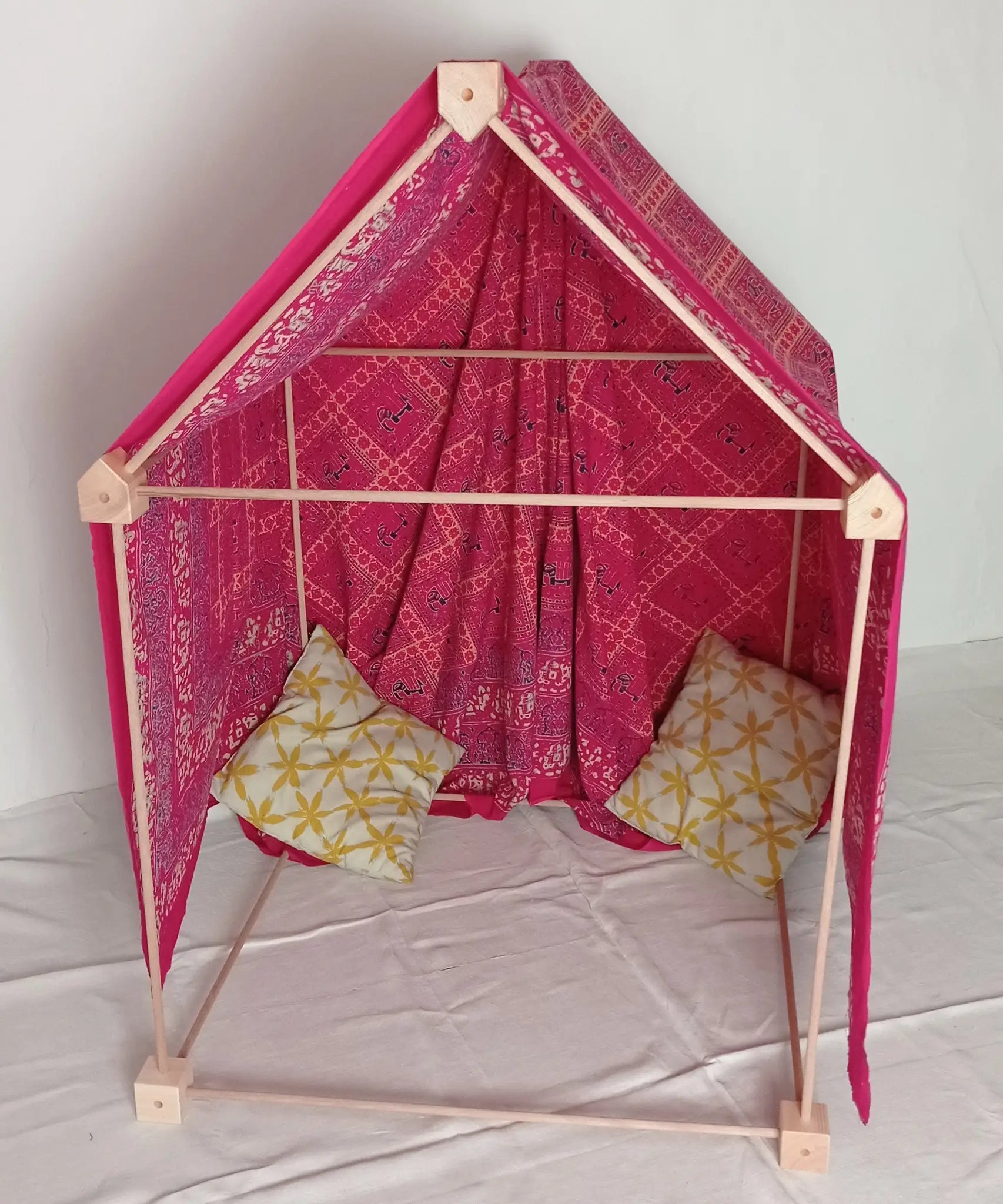 A cosy tent setup made from the Trigonos wooden big start set showing a decorated pink cloth on top of the frame and cushions inside