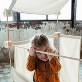 Child standing inside a tent showing wooden poles, blocks and white cotton sheets from the Trigonos kernel organic family constructions set
