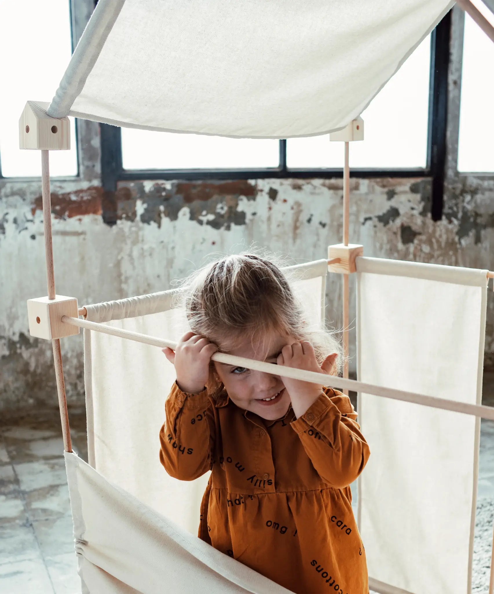 Child standing inside a tent showing wooden poles, blocks and white cotton sheets from the Trigonos kernel organic family constructions set