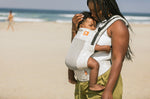 Close up of woman on a beach wearing the Tula beige Coast Isle FTG baby carrier