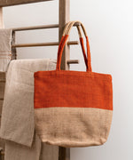 The Turtle Bags hand woven fair trade jute tote bag in an orange paprika colour block design, hanging on a wooden drying rack