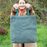 A woman holding up the Turtle Bags hand woven jute tote bag, in a teal stripe design with medium length handles