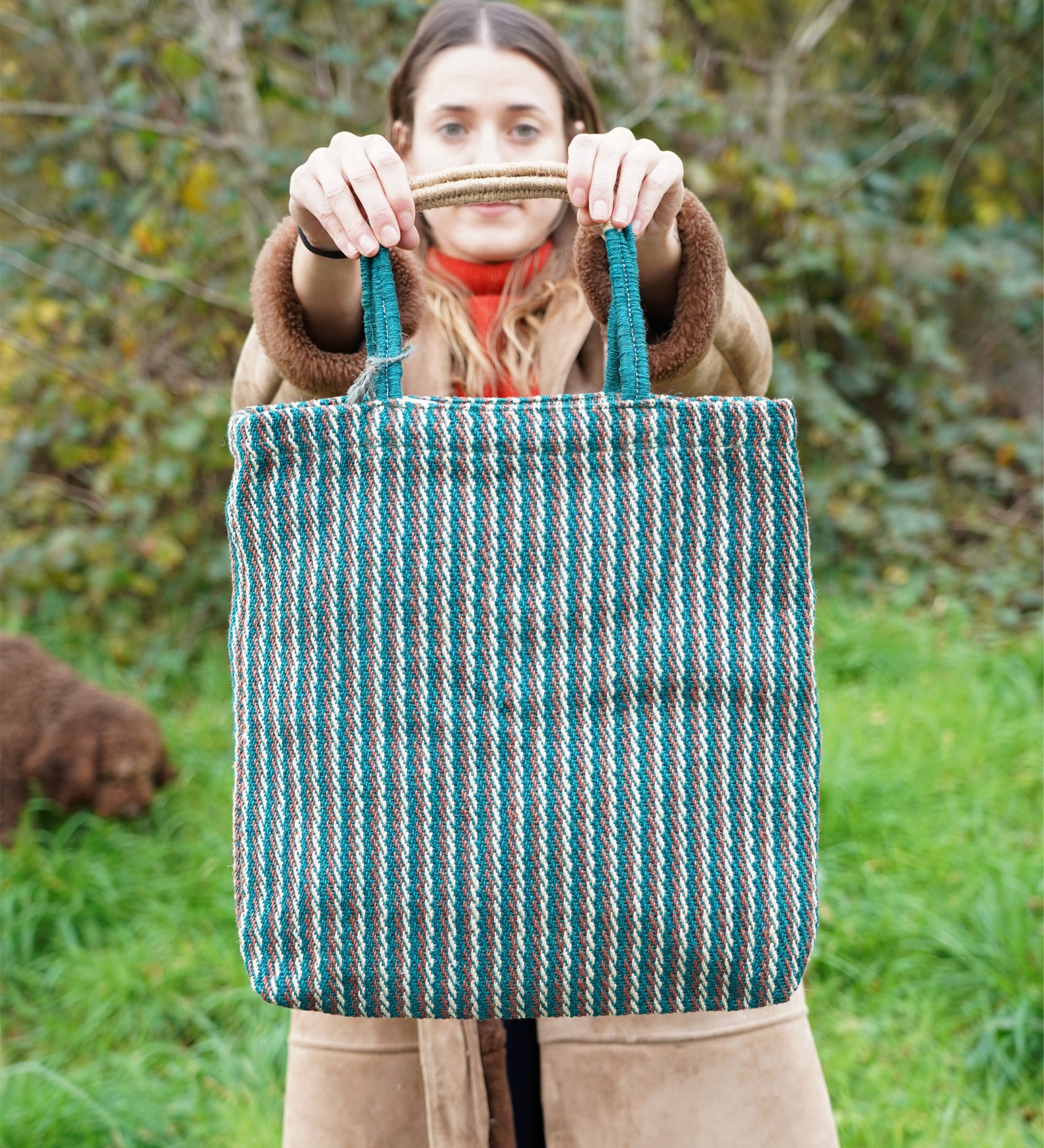 A woman holding up the Turtle Bags hand woven jute tote bag, in a teal stripe design with medium length handles