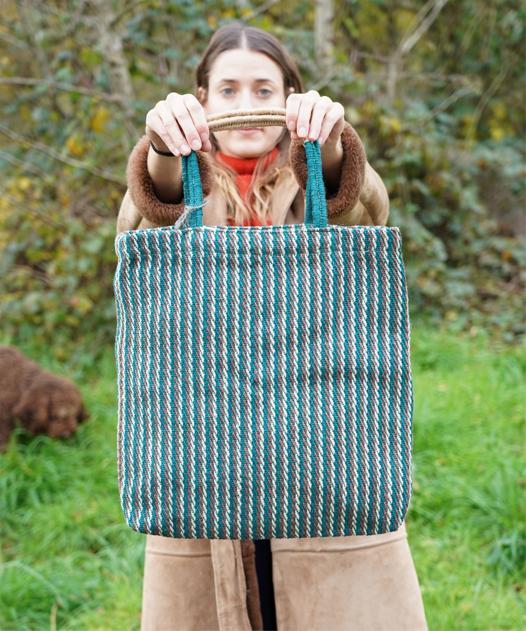 A woman holding up the Turtle Bags hand woven jute tote bag, in a teal stripe design with medium length handles