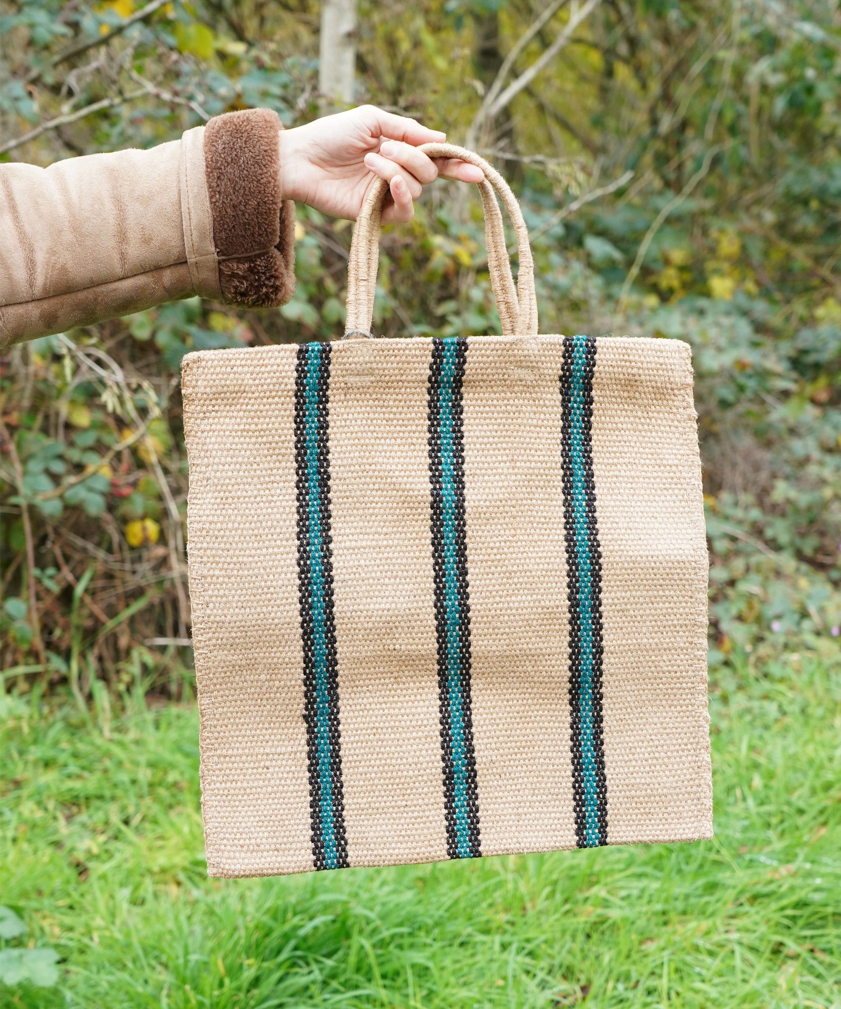 A hand holding out The Turtle Bags hand woven jute tote bag in a green and black stripe design