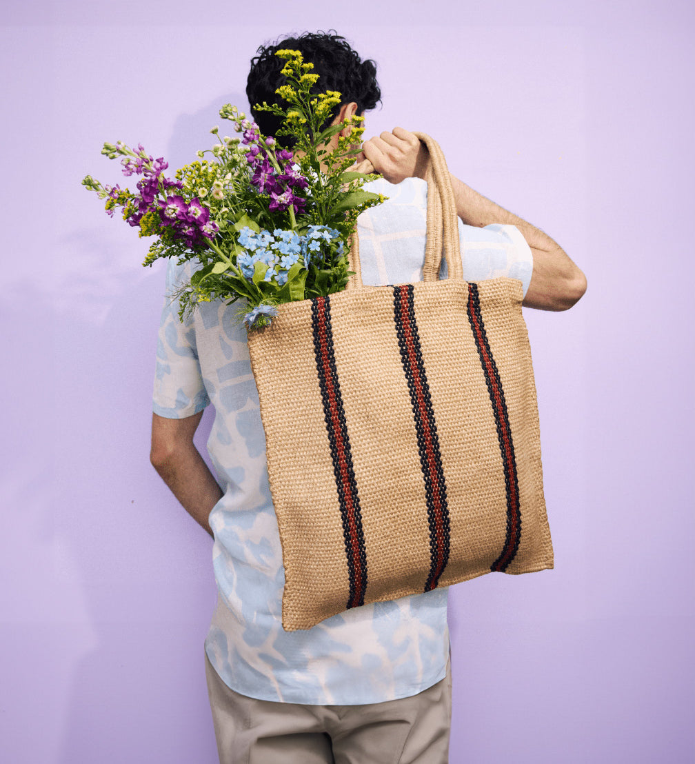 The back of a man carrying a Turtle Bags hand woven jute shopper tote bag in a red stripe design over his shoulder, with a bouquet of flowers inside