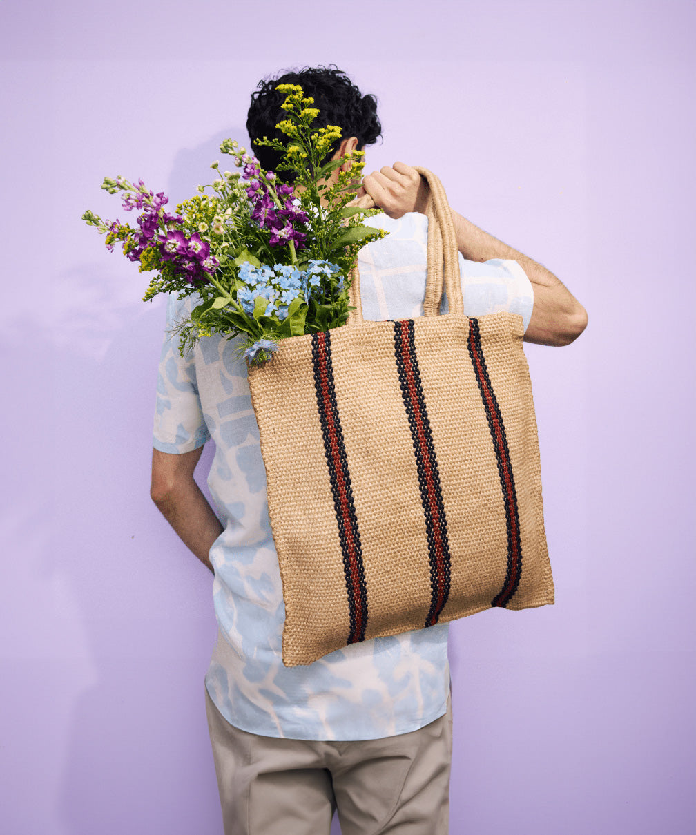 The back of a man carrying a Turtle Bags hand woven jute shopper tote bag in a red stripe design over his shoulder, with a bouquet of flowers inside