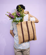 The back of a man carrying a Turtle Bags hand woven jute shopper tote bag in a red stripe design over his shoulder, with a bouquet of flowers inside