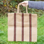 A hand holding The Turtle Bags hand woven jute tote bag with short handles in a red and black stripe design
