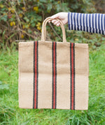 A hand holding The Turtle Bags hand woven jute tote bag with short handles in a red and black stripe design