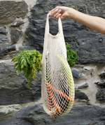 A hand holding a Turtle Bags fair trade and organic cotton string grocery shopping bag, with carrots and other vegetables inside.