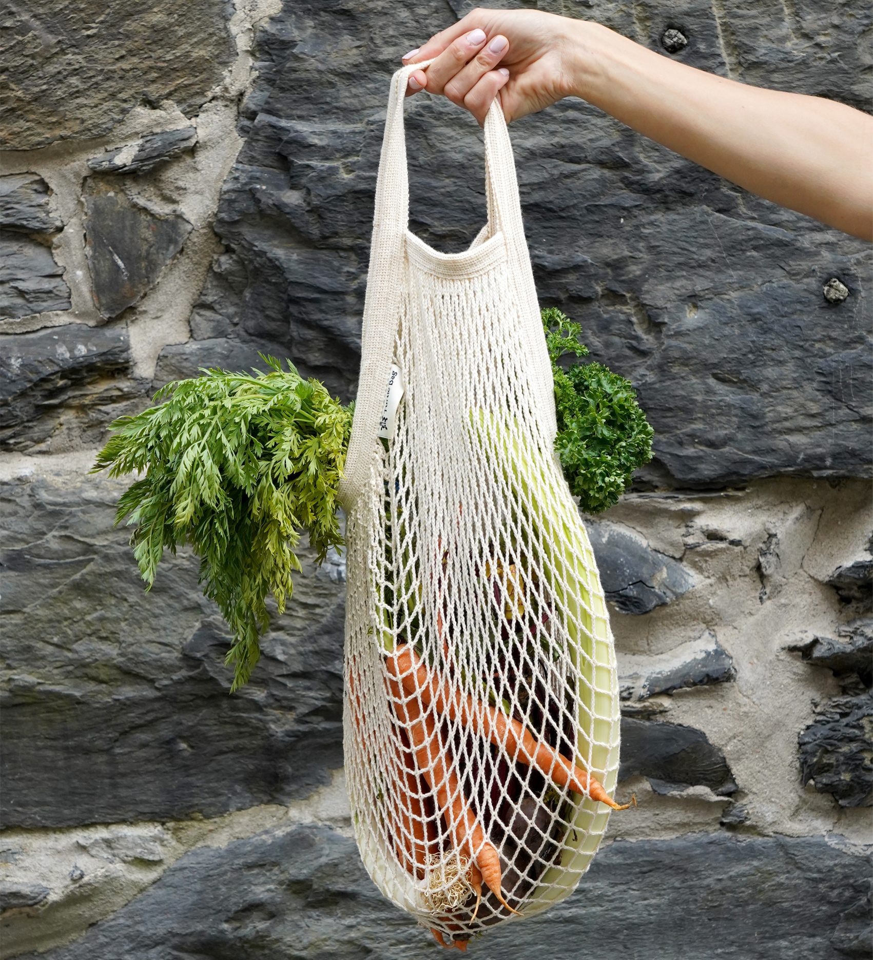 A hand holding a Turtle Bags fair trade and organic cotton string grocery shopping bag, with carrots and other vegetables inside.