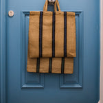 Turtle Bags Blue and Black Stripey Bag. A natural jute bag with short handles and 3 vertical blue and black stripes. Hanging off a central handle on a royal blue door with jute doormat on the floor