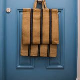 Turtle Bags Blue and Black Stripey Bag. A natural jute bag with short handles and 3 vertical blue and black stripes. Hanging off a central handle on a royal blue door with jute doormat on the floor