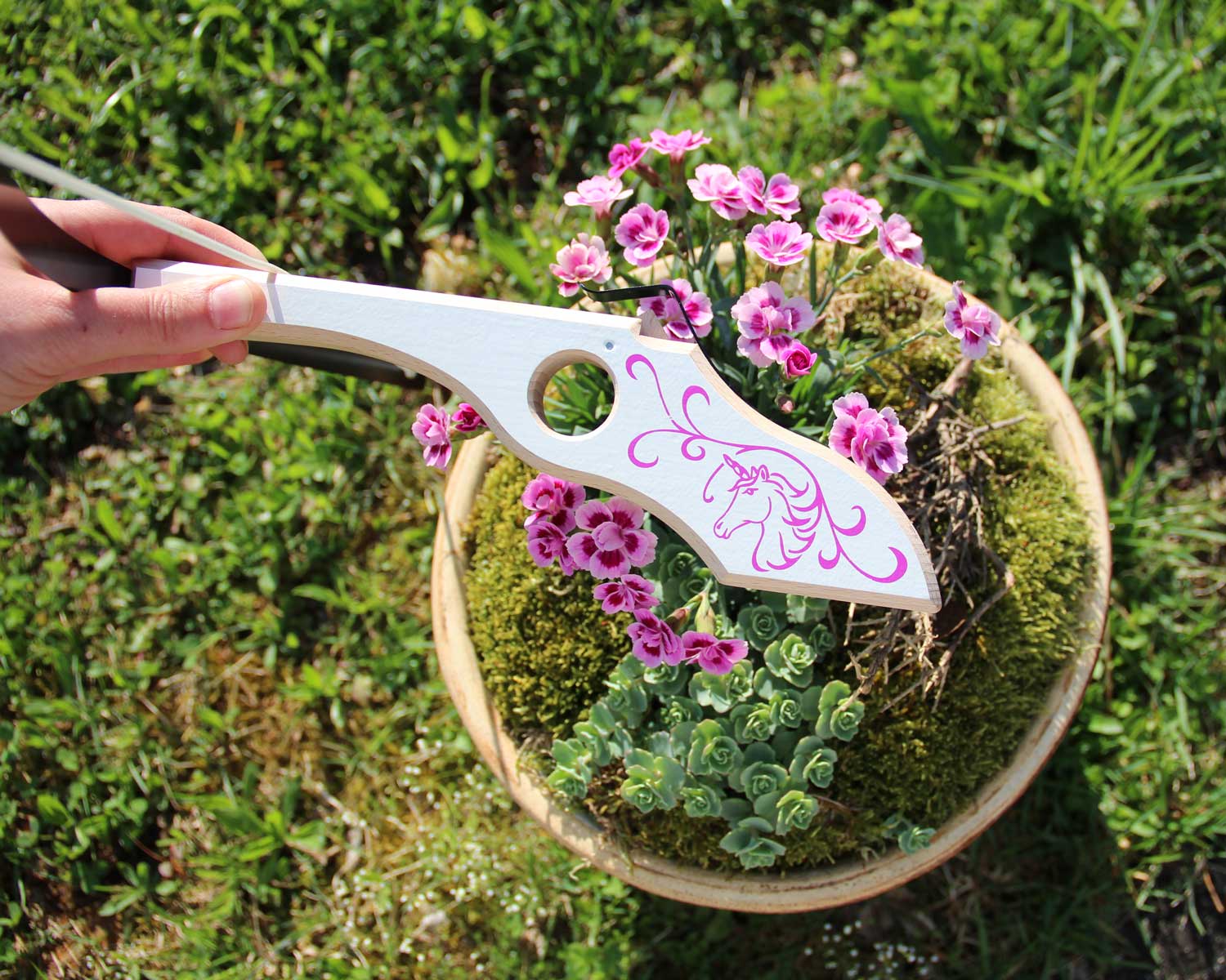 Person holding the Vah Arabella Unicorn Wooden Crossbow showing the design on the side with grass and flowers in the background