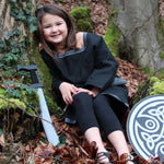 A child wearing a Vah Black Aragon child's Tunic, with a round shield and sword placed besides her on the floor. The child is sitting down on a forest.