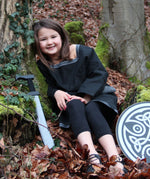 A child wearing a Vah Black Aragon child's Tunic, with a round shield and sword placed besides her on the floor. The child is sitting down on a forest.