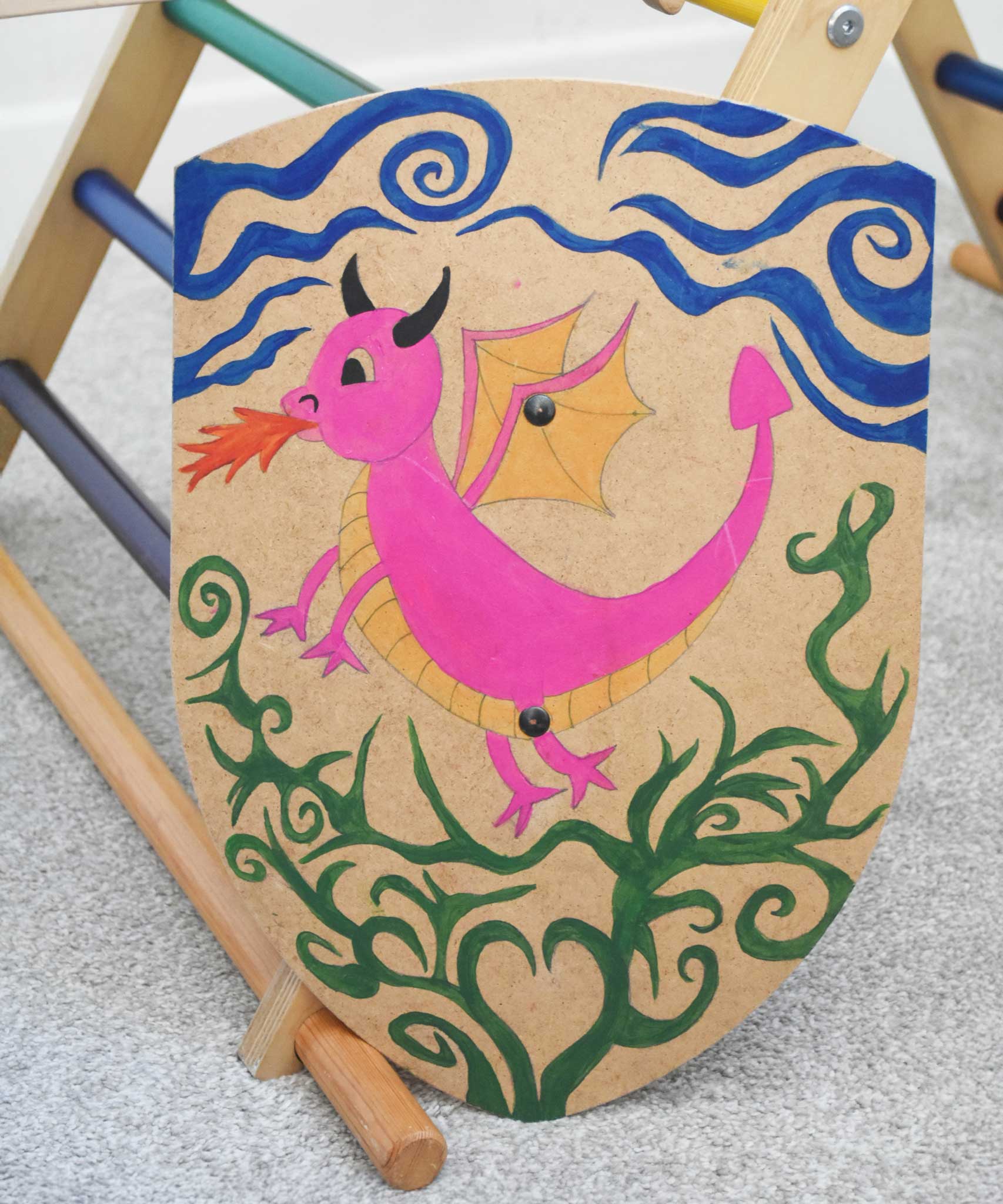 A VAH DIY shield hand painted with a pink dragon. The Shield has been placed resting on the side of a Trimblimb climbing frame. 