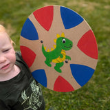 A young child holding up a hand painted VAH circle shaped DIY wooden shield. The shield has a hand-drawn hand-painted green dragon in the middle and blue and red coloured triangle shapes around the edges. 