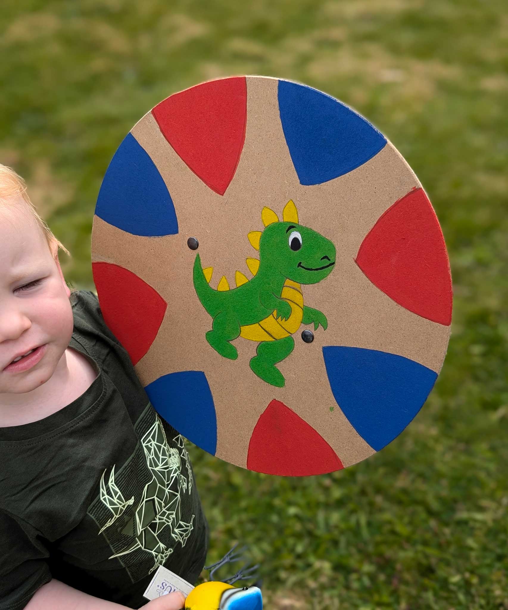 A young child holding up a hand painted VAH circle shaped DIY wooden shield. The shield has a hand-drawn hand-painted green dragon in the middle and blue and red coloured triangle shapes around the edges. 