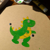 A circle shaped, blank VAH wooden shield with a dinosaur drawn in pencil and painted with green and yellow paint. A paint pallet and tubes of paint can be seen in the background.