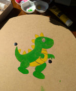 A circle shaped, blank VAH wooden shield with a dinosaur drawn in pencil and painted with green and yellow paint. A paint pallet and tubes of paint can be seen in the background.