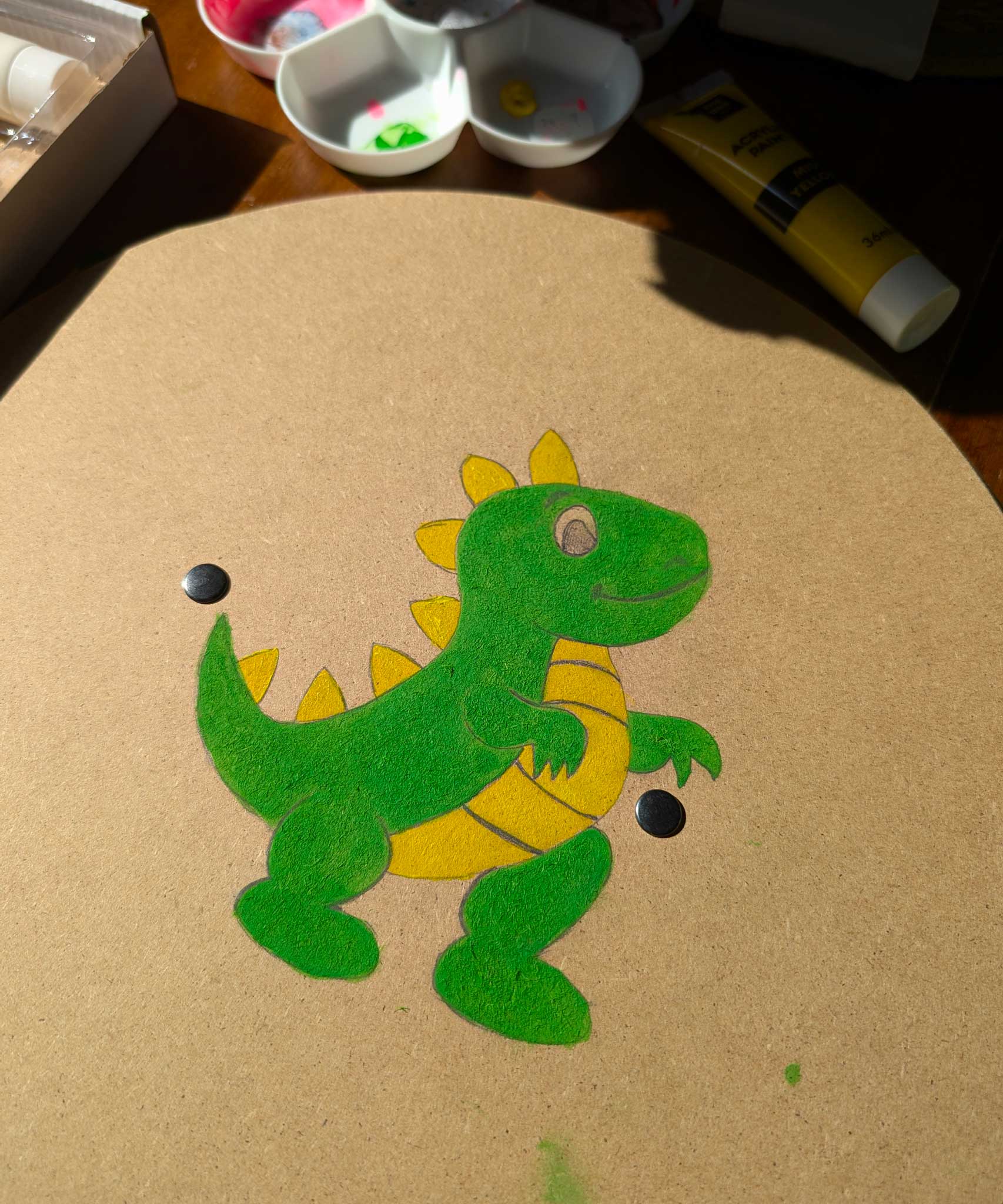 A circle shaped, blank VAH wooden shield with a dinosaur drawn in pencil and painted with green and yellow paint. A paint pallet and tubes of paint can be seen in the background.