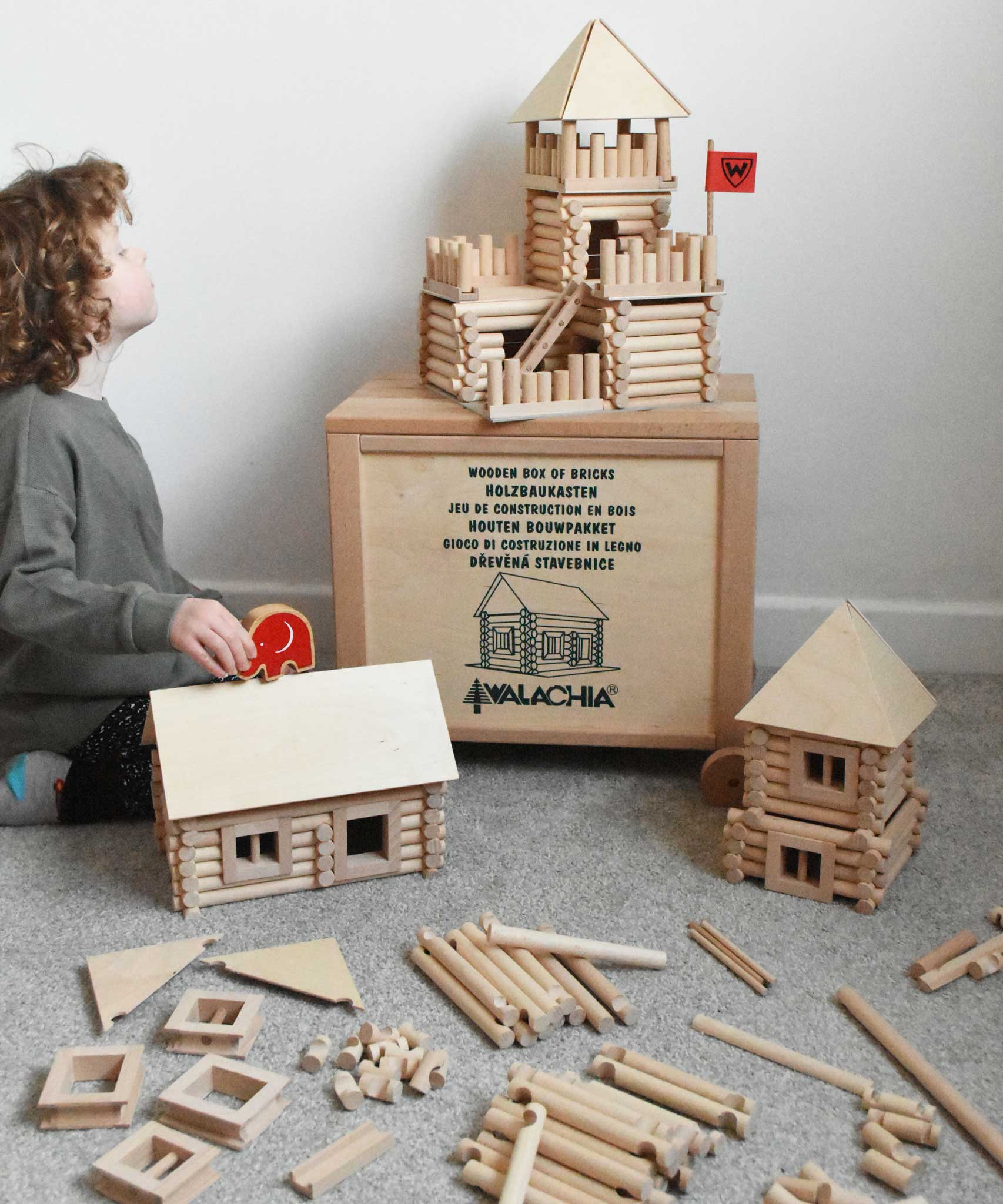 Walachia Vario Box & Fort Building Set 450 Pieces. The blocks are supplied in a large wooden storage case. There are three different builds around the wooden crate. A child in sitting besides the crate and is placing a Babipur elephant figure on the roof of a log cabin build.
This modular wooden building block system is perfect for those who enjoy a challenge. The sets are available in various sizes at Babipur. 