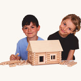 Two children smiling with a log cabin created using wooden blocks from the Walachia Vario Building Sets in front of them. This modular wooden building block system is perfect for those who enjoy a challenge. The sets are available in various sizes at Babipur.  
