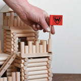 A close up of an adult's hand placing the the flag pole in place on a fort build created using the Walachia Vario Fort Building Set 194 Pieces. This modular wooden building block system is perfect for those who enjoy a challenge. The sets are available in various sizes at Babipur. 