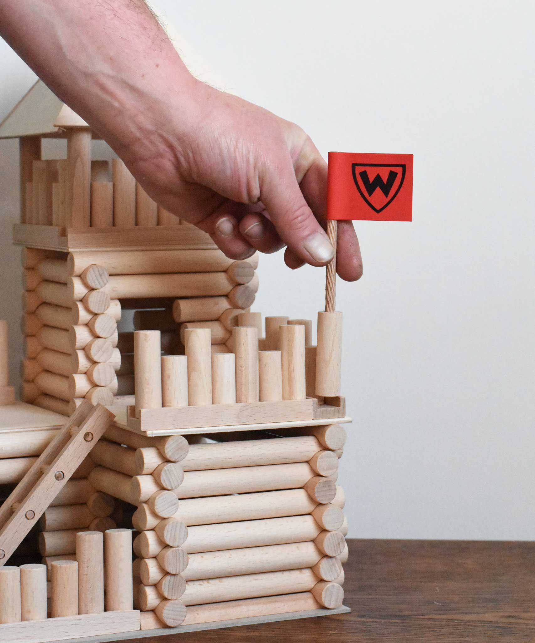 A close up of an adult's hand placing the the flag pole in place on a fort build created using the Walachia Vario Fort Building Set 194 Pieces. This modular wooden building block system is perfect for those who enjoy a challenge. The sets are available in various sizes at Babipur. 